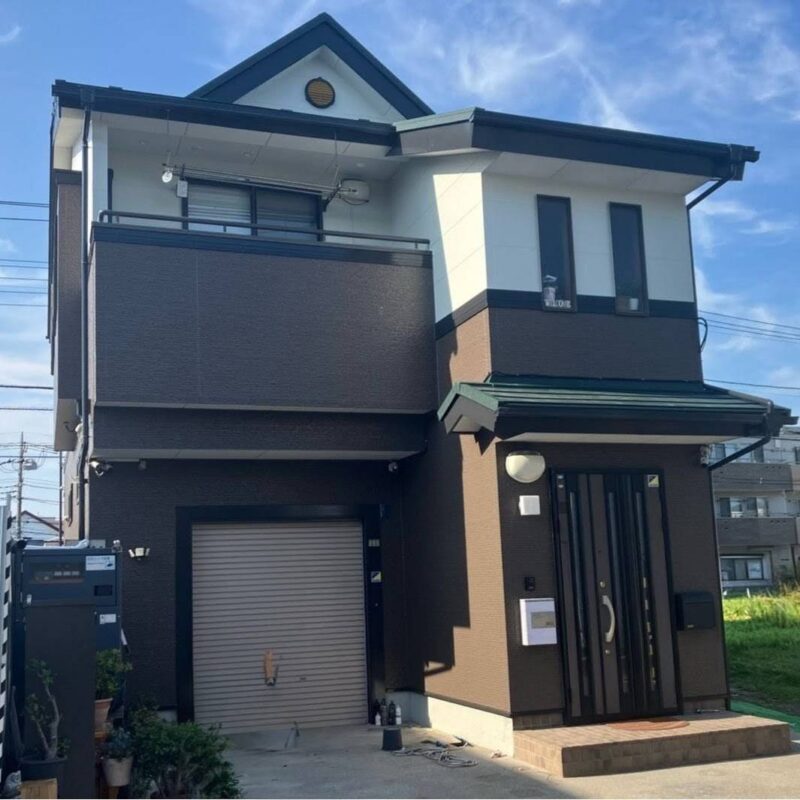 ブラウンの木目調外壁とホワイトのツートンカラーに塗り分けた、ガレージ付き3階建て住宅の施工後写真。晴天の下、落ち着いた配色が周囲の景観と調和しており、窓枠や破風板の濃いブラウンが全体をスタイリッシュに引き締めている。