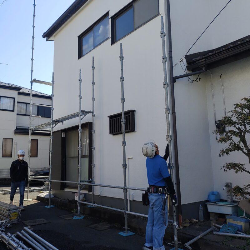 外壁塗装工事のために、2階建て住宅の白いモルタル外壁の周囲にしっかりと組み立てられた金属製の足場。