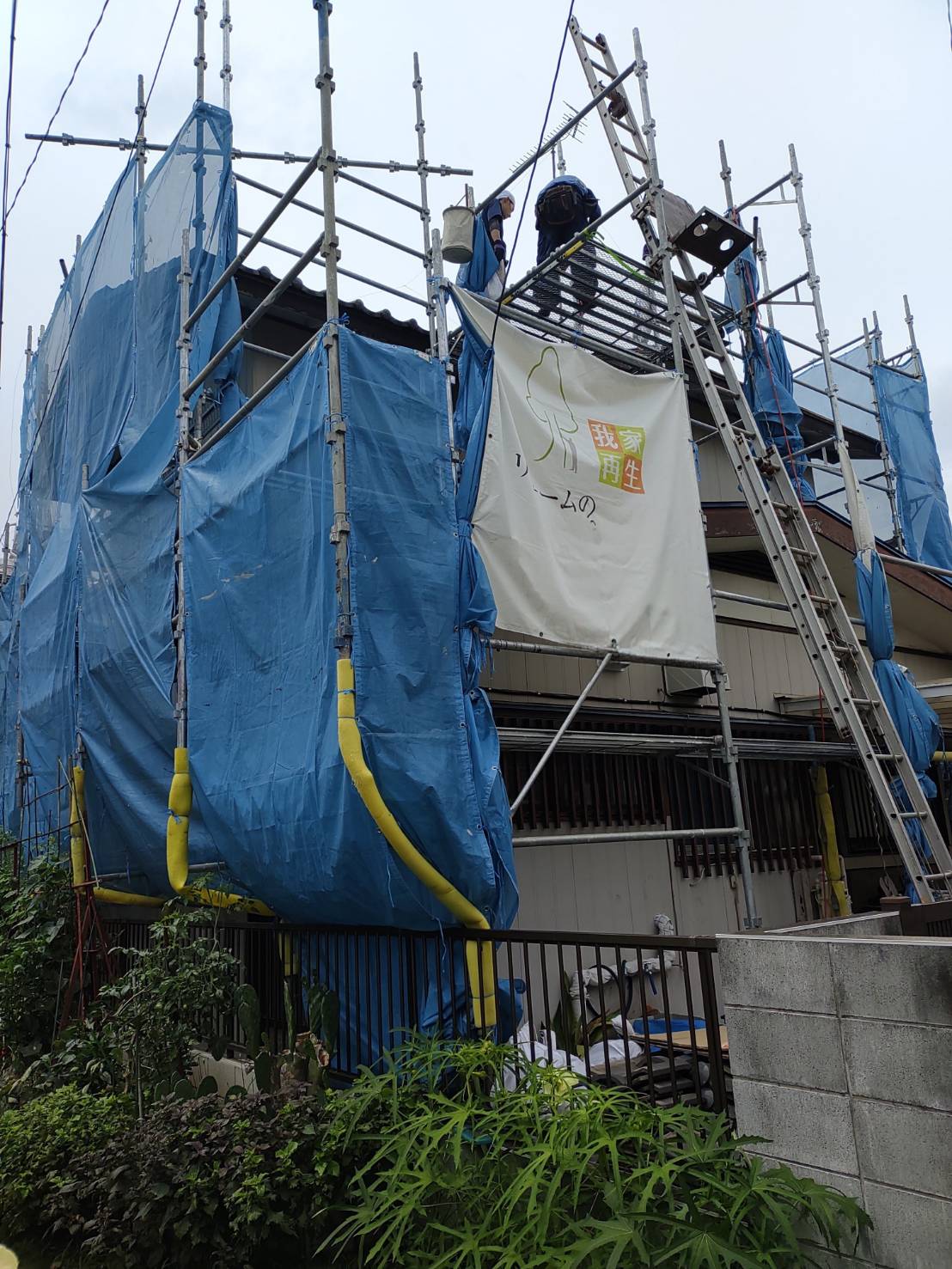 川口市での外壁リフォーム・屋根葺き替え工事にともなう足場仮設工事。住宅の周囲に金属製の足場を組み、安全確保と近隣への飛散防止のためのブルーシート(養生ネット)を張った状態。