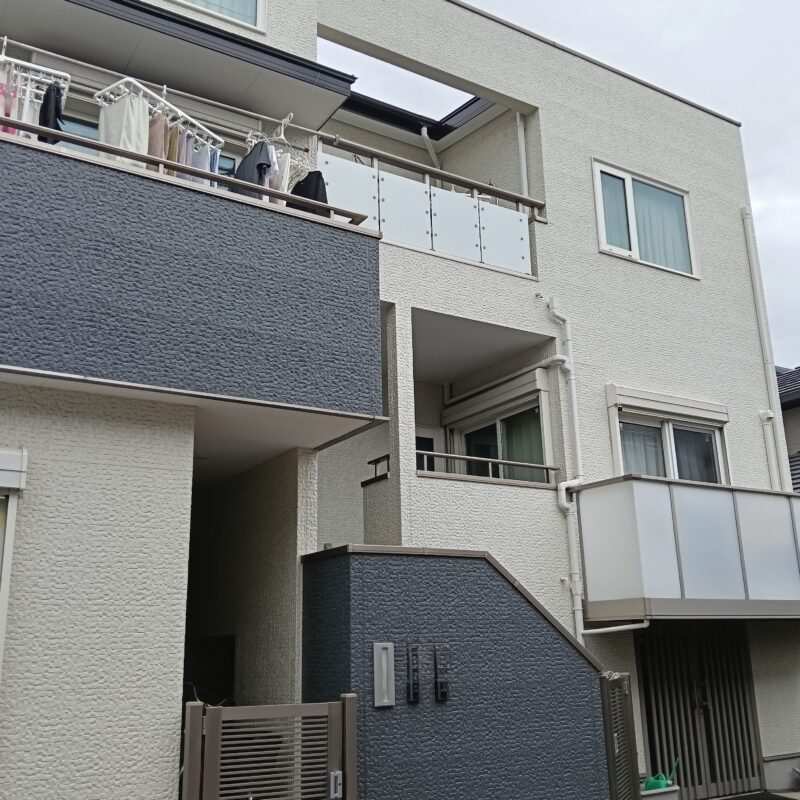 川口市でのデザイン住宅の外装リフォーム完了写真。ベースの白い外壁には雨だれに強い低汚染塗装を施し、バルコニーや1階門袖の石積み調アクセント壁を深いダークグレーで塗り分けたモダンな多層構造・3階建て住宅の外観。屋根の葺き替えも行われ、曇り空のもとで新築のようにシャープでメリハリのある美観を取り戻した施工事例。