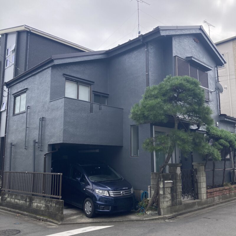 川口市での外装リフォーム完了写真。屋根を軽量な最新の屋根材へと葺き替え、外壁には重重感と高級感のあるダークネイビー（チャコールグレー）の塗装を施した和モダンな2階建て住宅の外観。手前には大切に手入れされた立派な松の木と風情ある門構えがあり、ガレージに紺色のミニバンが駐車されている、伝統と現代的デザインが美しく調和した施工事例。