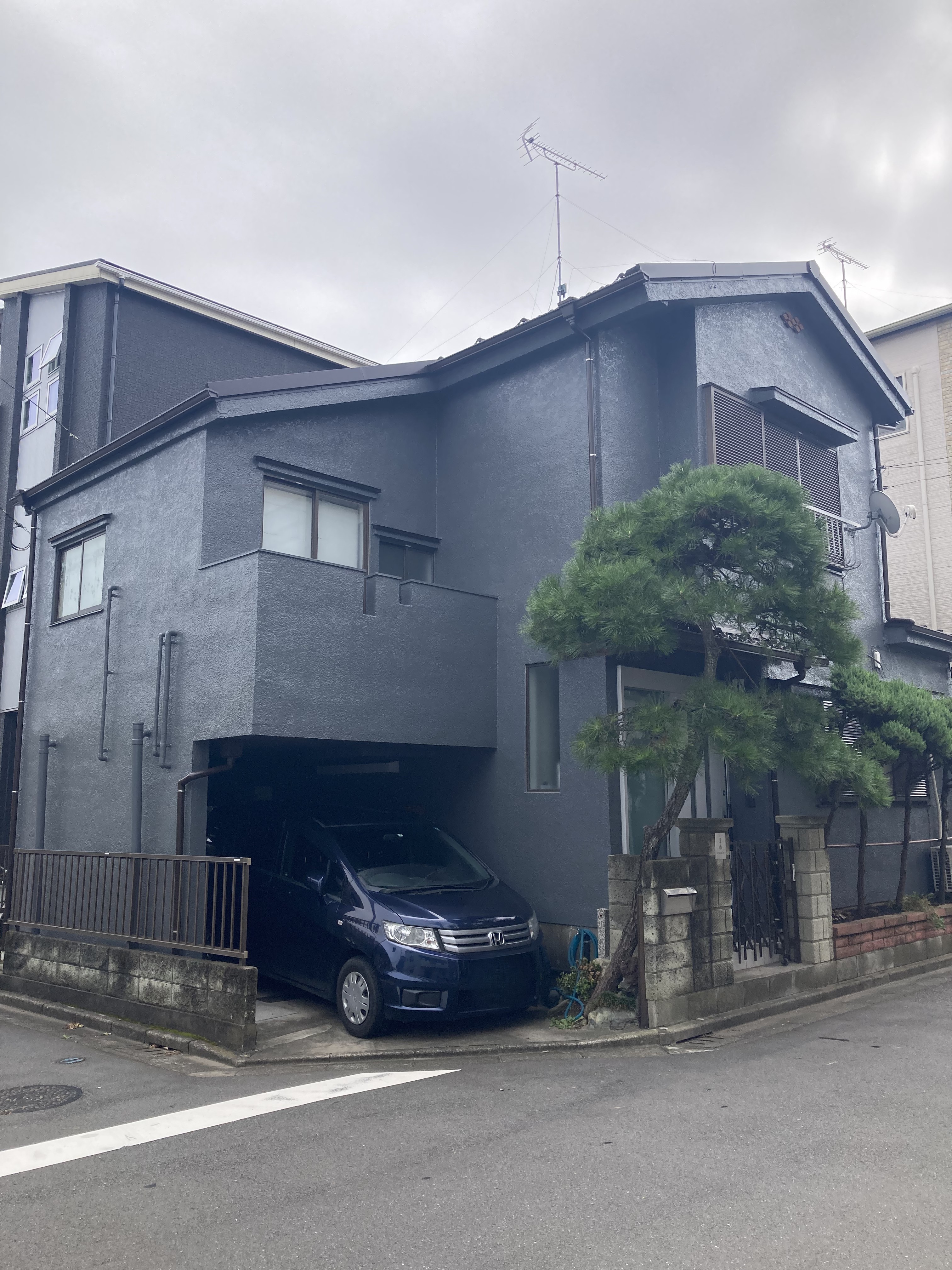川口市での外装リフォーム完了写真。屋根を軽量な最新の屋根材へと葺き替え、外壁には重重感と高級感のあるダークネイビー（チャコールグレー）の塗装を施した和モダンな2階建て住宅の外観。手前には大切に手入れされた立派な松の木と風情ある門構えがあり、ガレージに紺色のミニバンが駐車されている、伝統と現代的デザインが美しく調和した施工事例。