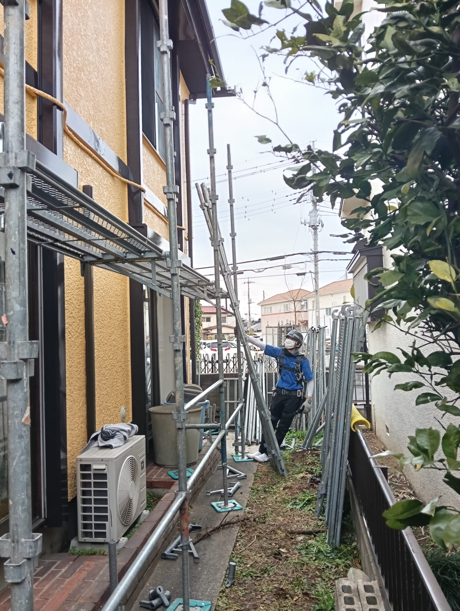 草加市の戸建て外壁塗装工事現場にて、全ての塗装と検査が完了した後、ヘルメットと安全帯を着用した足場職人が、エアコン室外機や庭木に配慮しながら犬走りで慎重に鉄骨を解体している様子