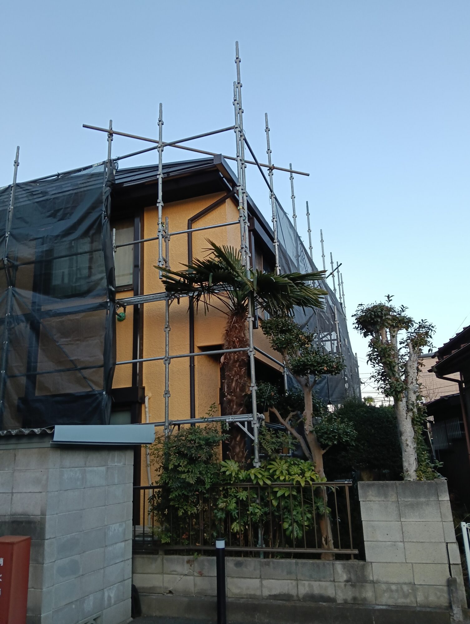 草加市の戸建て外壁塗装工事にて、すべての塗装工程が美しく塗り上がり、足場解体前の最終検査を控えたお住まいの足場越し・メッシュシート越しの外観写真