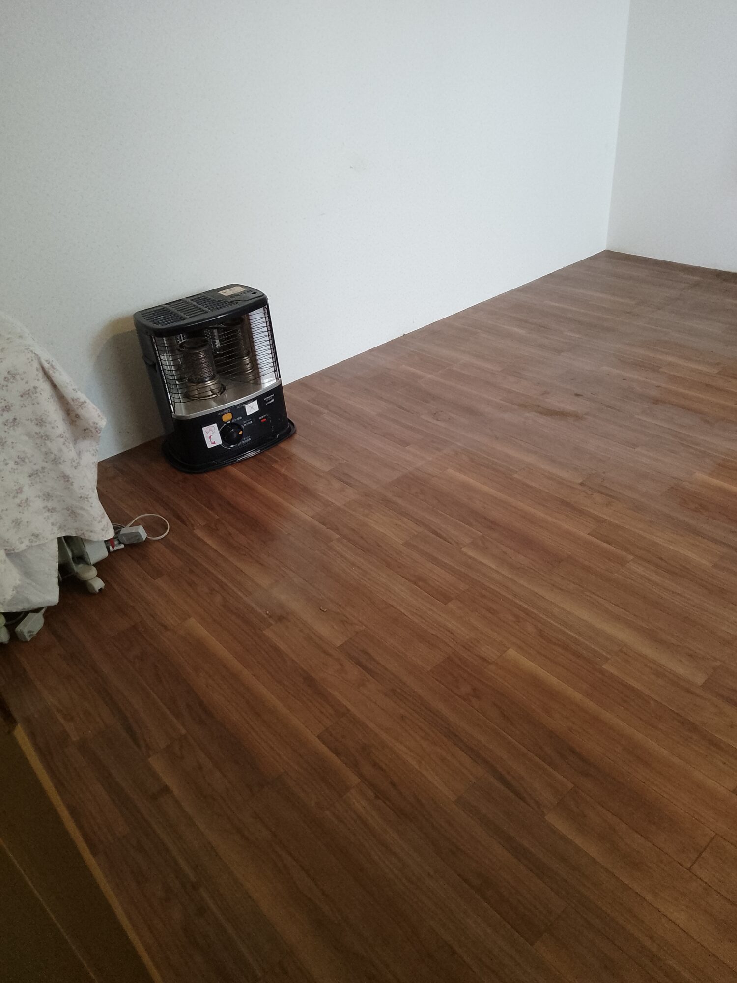 草加市K邸の内装リフォーム施工前(Before)写真。経年により全体的にくすんで白っぽくなった壁紙(クロス)と、やや濃いブラウンの床材の様子。天井からはシンプルな裸電球のペンダントライトが下がり、正面の窓にはベージュのカーテンが閉められているリフォーム前の居室空間。