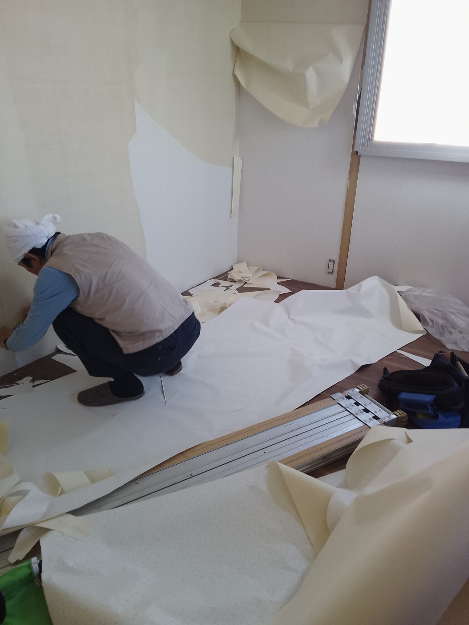 草加市K邸での内装リフォームにおける、クロス(壁紙)張替えの施工中の様子。脚立にのった内装職人が、天井近くの古い壁紙を丁寧に手で剥がしている。白く新しい壁面と、剥がされた後に露出したベージュ色の下地が対比となっており、足元には道具類やバケツが置かれている、リフォームの丁寧な下地準備の工程が伝わる作業風景。