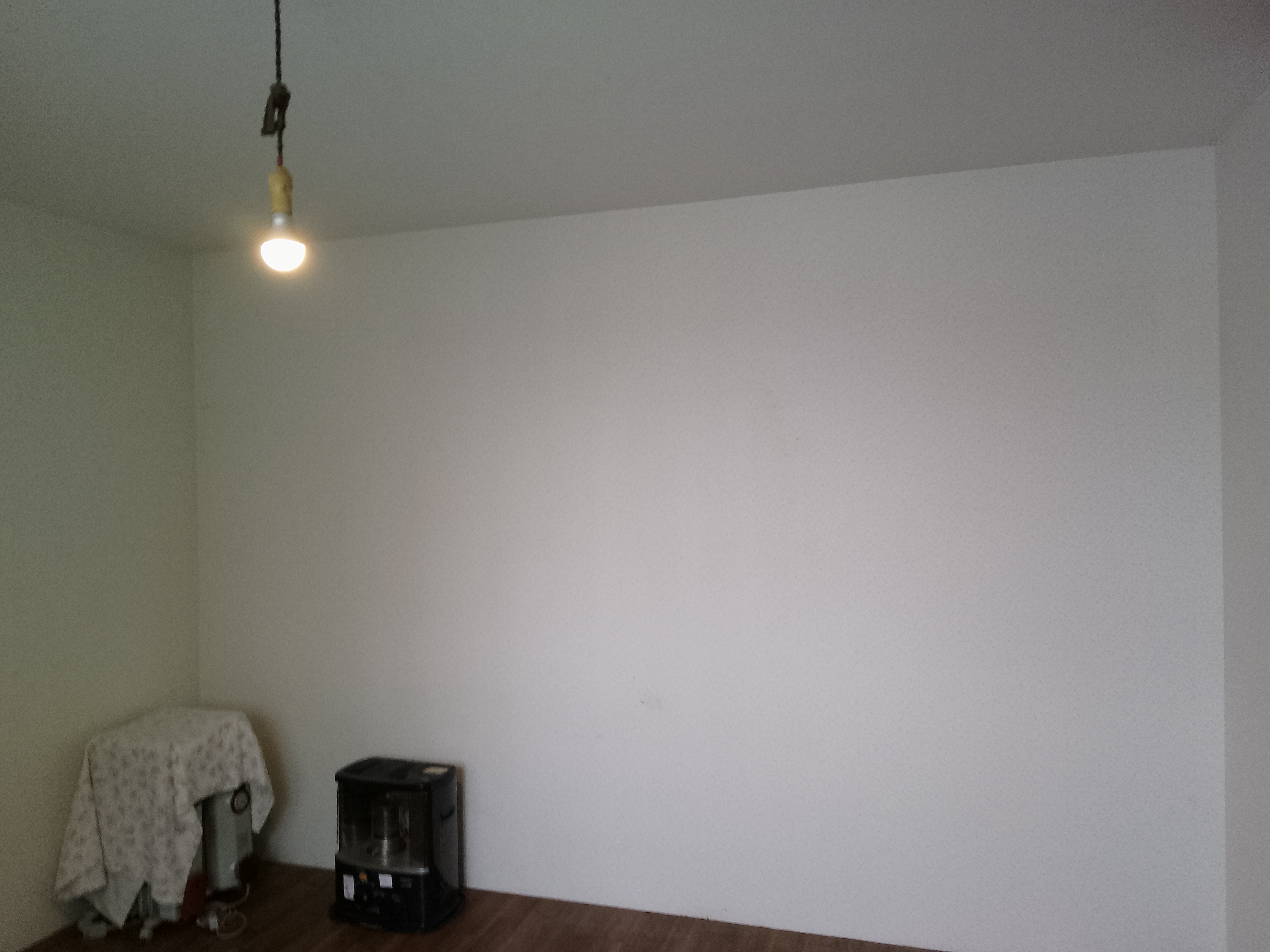 草加市K邸の内装リフォーム施工前(Before)写真。経年により全体的にくすんで白っぽくなった壁紙(クロス)と、やや濃いブラウンの床材の様子。天井からはシンプルな裸電球のペンダントライトが下がり、正面の窓にはベージュのカーテンが閉められているリフォーム前の居室空間。