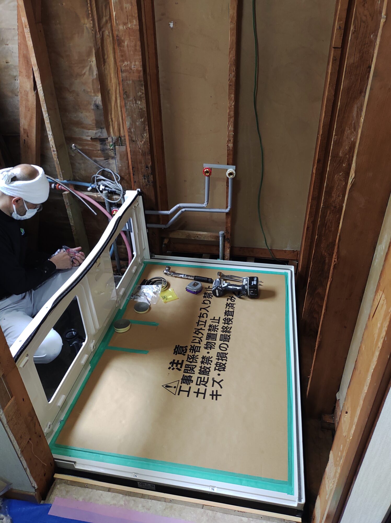 草加市のお風呂リフォーム現場にて、新設したユニットバスの床パネルに養生ダンボールを敷き詰め、土足厳禁の徹底管理のもとで本体パネルやドア枠の組み立て作業を行う職人の写真
