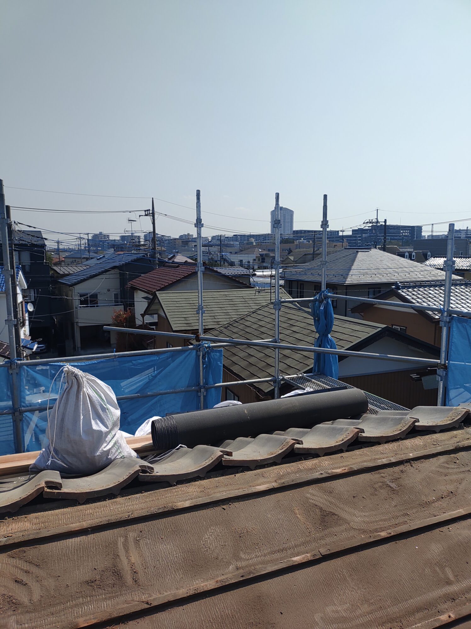 草加市の戸建て屋根葺き替えリフォーム現場にて、瓦撤去後の下地に敷設するための新しい防水シート(ルーフィング)のロールが置かれた、安全足場に囲まれた高所作業中の写真
