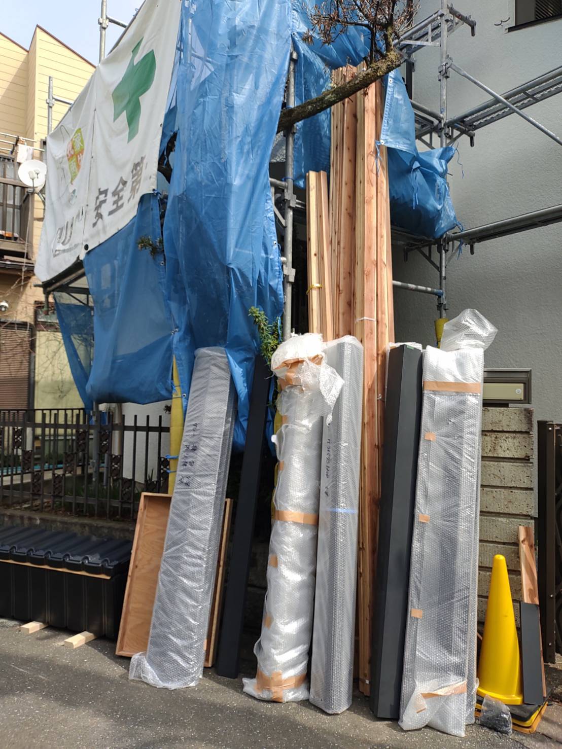 草加市の屋根葺き替え工事現場にて、搬入された新しいガルバリウム鋼板の金属屋根資材や下地木材が、傷防止のため緩衝材で丁寧に梱包され、安全足場の前に美しく並べられた写真
