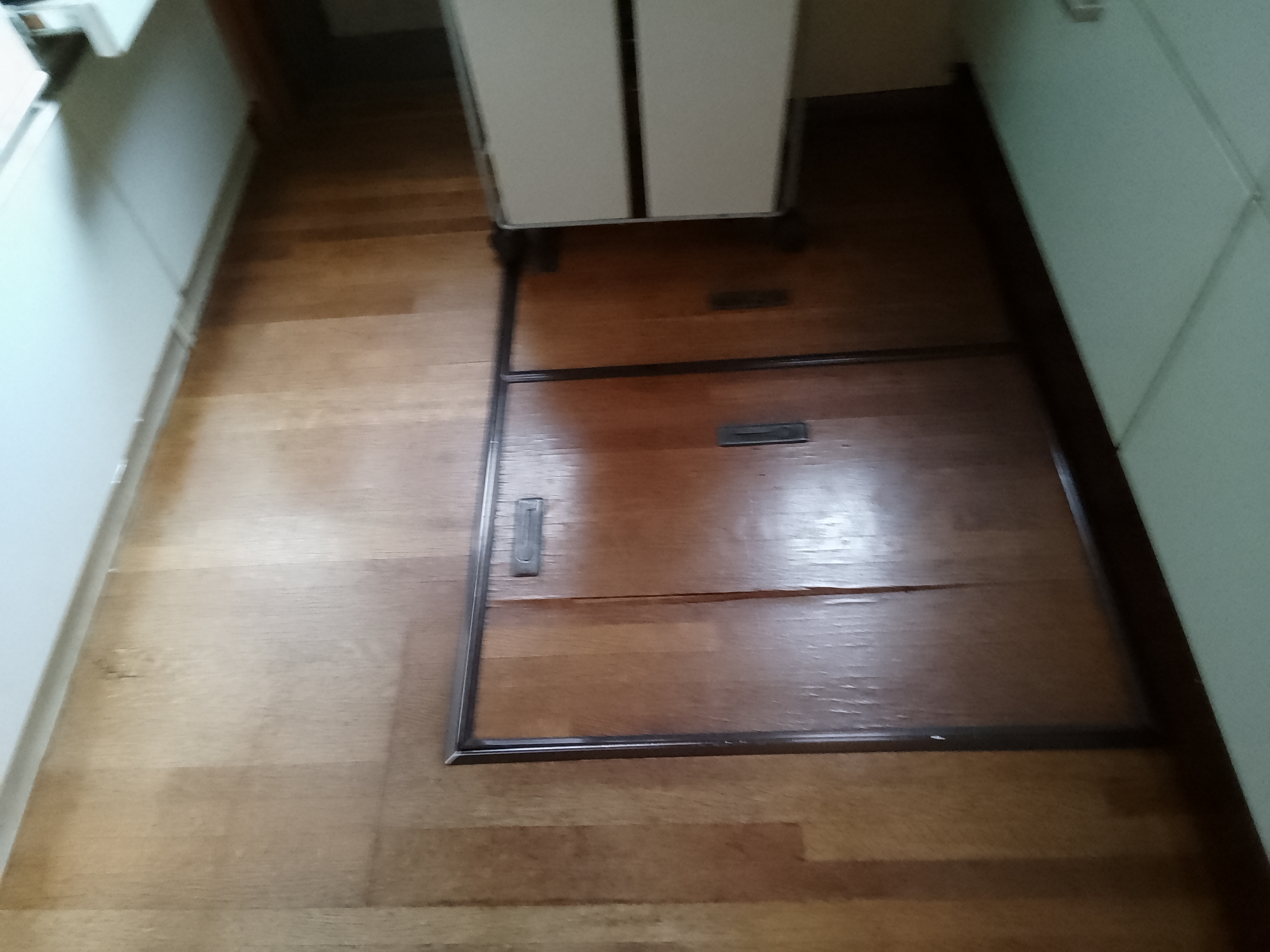 草加市のキッチン床リフォーム現場にて、古いフローリングの上に新しい床材を重ねて張る上張り工事を行う前の、床下収納（点検口）がある状態を撮影した着工前の写真